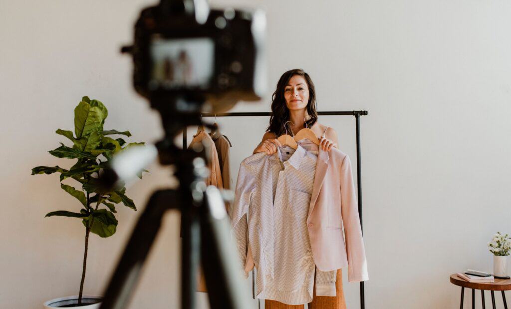 Una mujer sostiene dos chaquetas frente a una cámara colocada sobre un trípode, con un perchero y una planta al fondo, y comparte sus conocimientos sobre moda. Isabel Restrepo, conocida por ser mentora de los entusiastas de la moda de Miami, combina estilo y consejos prácticos sin esfuerzo.