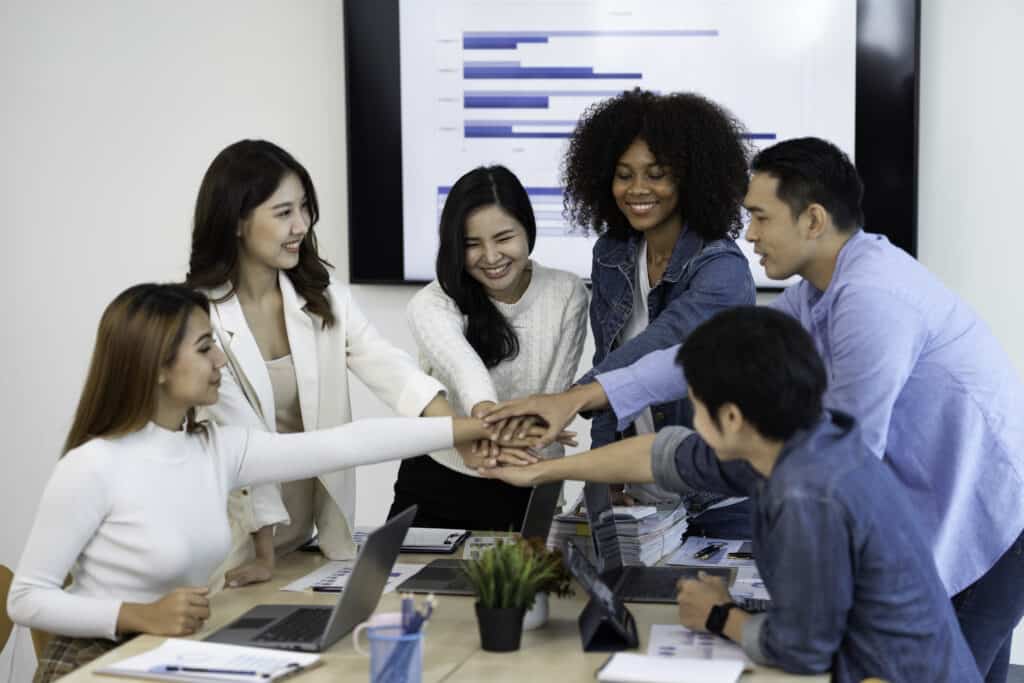 Un grupo diverso de seis colegas se encuentran de pie alrededor de una mesa en una oficina, colocando sus manos en el centro en un gesto de trabajo en equipo. Sobre la mesa se ven gráficos y documentos, y en una pantalla detrás de ellos se muestra la leyenda "Mentoring Miami".