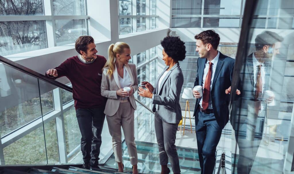 Cuatro profesionales de negocios, tres mujeres y un hombre, están de pie en una escalera de un moderno edificio de oficinas, conversando y sosteniendo tazas de café. Entre ellos se encuentra Isabel Restrepo, una destacada oradora de Miami conocida por su trabajo de mentoría en la ciudad.