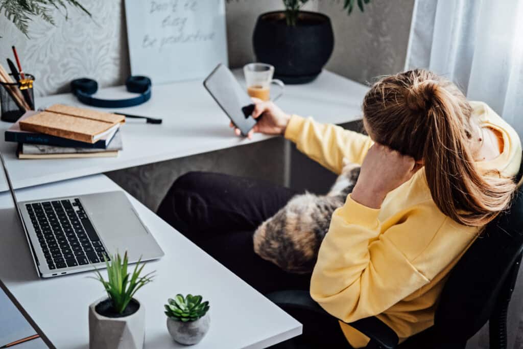 Persona con sudadera amarilla sentada en un escritorio con una computadora portátil, libros y plantas, sosteniendo una tableta mientras un gato se sienta en su regazo. Isabel Restrepo, conocida por Mentoring Miami, podría estar preparando su próxima charla.