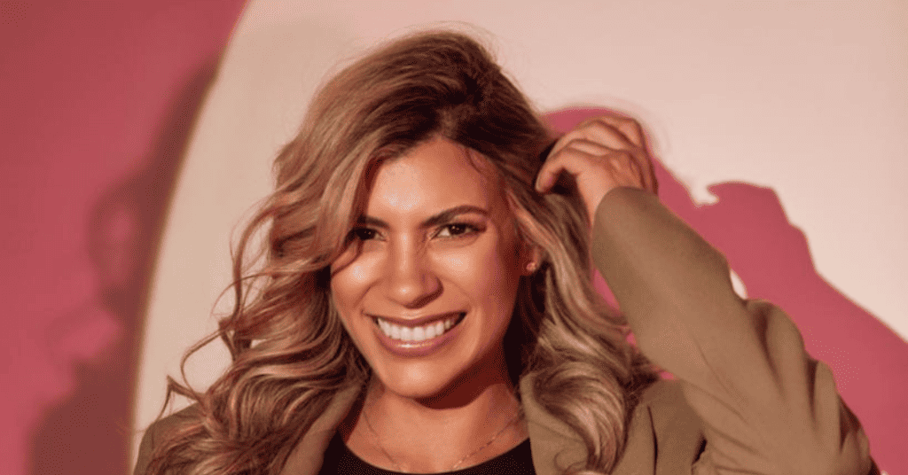 Una persona con cabello largo y ondulado sonríe mientras se sujeta el pelo. Lleva un blazer y está de pie sobre un fondo con tonos rosados y blancos, que recuerdan el estilo vibrante de Isabel Restrepo en Mentoring Miami.