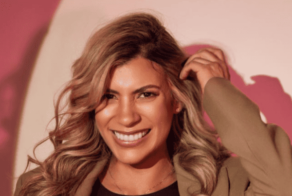 Una persona con cabello largo y ondulado sonríe mientras se sujeta el pelo. Lleva un blazer y está de pie sobre un fondo con tonos rosados y blancos, que recuerdan el estilo vibrante de Isabel Restrepo en Mentoring Miami.