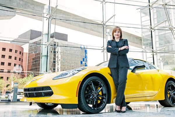 Una persona vestida de negocios se encuentra de pie con confianza y los brazos cruzados junto a un automóvil deportivo amarillo dentro de un edificio moderno con grandes ventanales y un telón de fondo urbano de Miami, encarnando el espíritu dinámico de Isabel Restrepo.