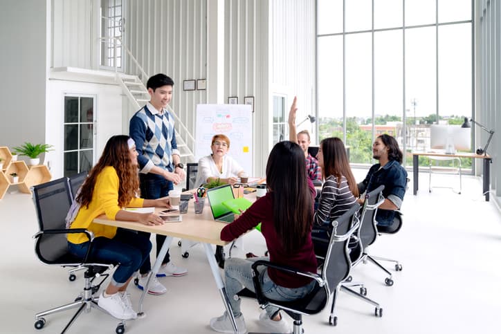 Un grupo de siete personas se reúnen alrededor de una mesa en una oficina moderna y participan en una discusión sobre habilidades de venta. Una persona está de pie y habla, mientras que las demás están sentadas y escuchan atentamente.