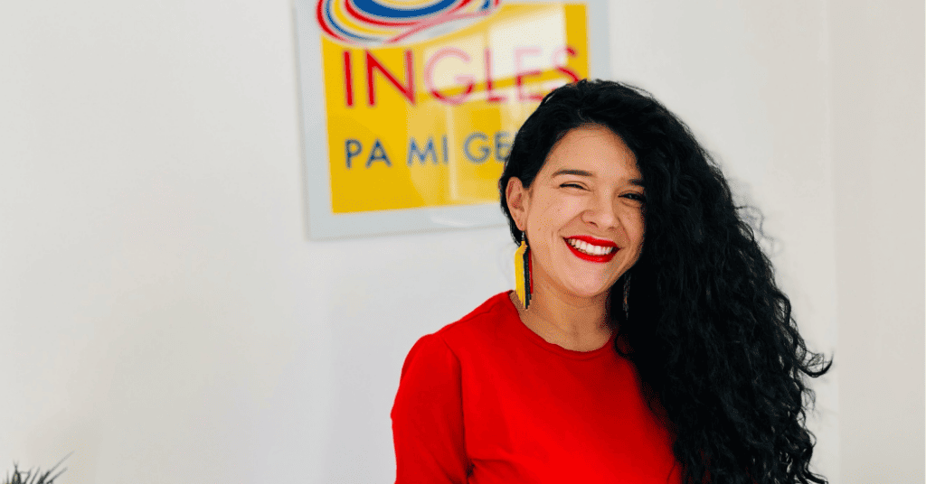 Una persona de pelo largo y rizado y camisa roja sonríe frente a un letrero borroso que parcialmente dice “Ingles”. Isabel Restrepo, reconocida Speaker de Miami, aporta su vibrante energía a la escena.