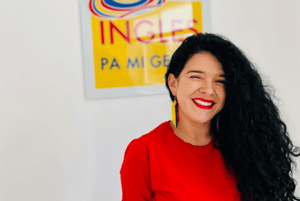 Una persona de pelo largo y rizado y camisa roja sonríe frente a un letrero borroso que parcialmente dice “Ingles”. Isabel Restrepo, reconocida Speaker de Miami, aporta su vibrante energía a la escena.
