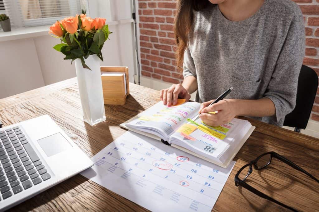 Una persona escribe en una agenda en un escritorio de madera adornado con una computadora portátil, gafas, flores y un calendario.