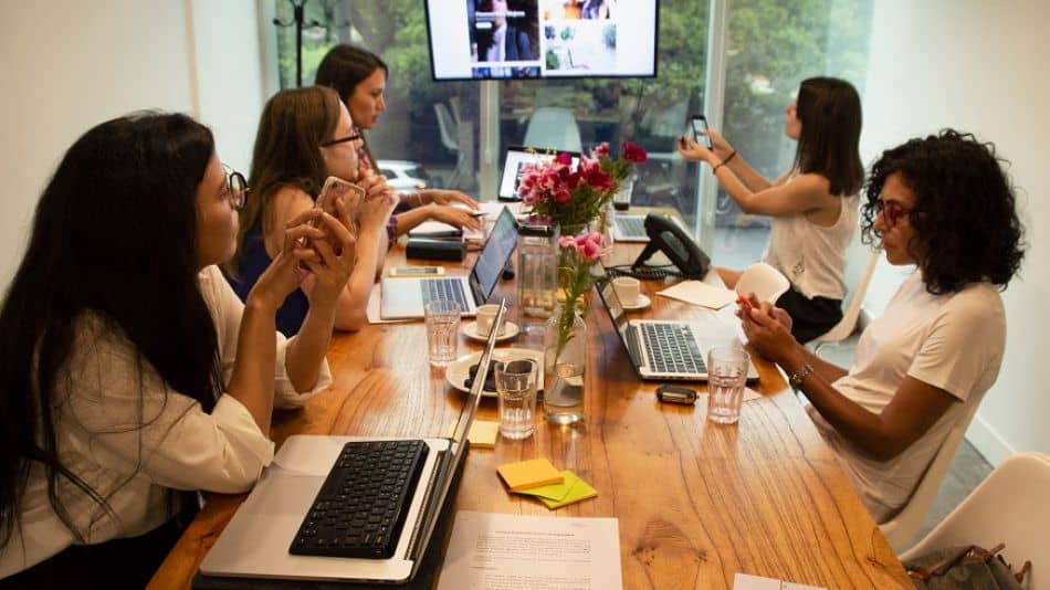 Cinco personas trabajan alrededor de una mesa en una oficina, usando computadoras portátiles y teléfonos inteligentes. La habitación tiene una gran ventana con vista a los árboles. Una pantalla en la pared muestra imágenes. Claramente, una reunión o sesión colaborativa con mujeres exitosas a la cabeza.