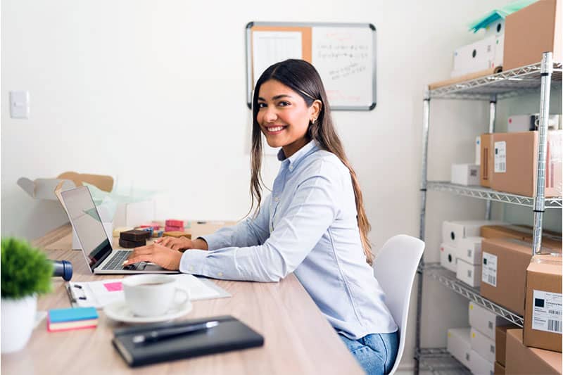 Una mujer está sentada en un escritorio con una computadora portátil y sonríe a la cámara. Detrás de ella, hay estantes con cajas y suministros para envíos. En la pared hay una pizarra con notas sobre habilidades de venta.