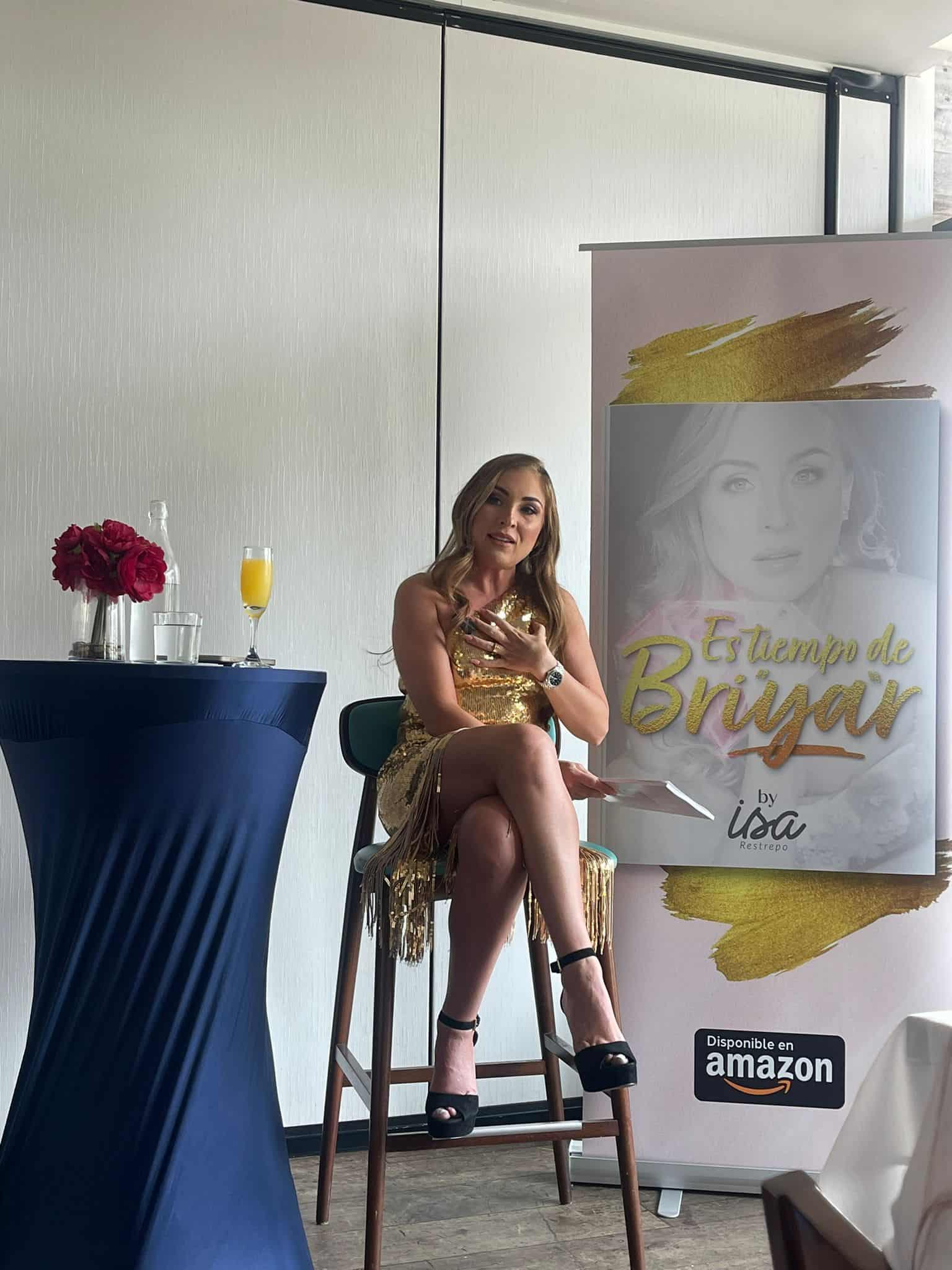 Una mujer con un vestido dorado está sentada en una silla alta junto a una mesa con flores rojas y una bebida. A su lado hay una pancarta que promociona un libro titulado "Es Tiempo de BiYAr" de Isa, disponible en Amazon.