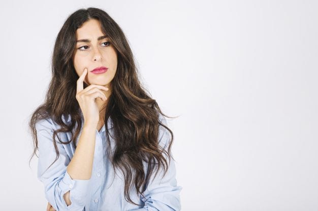 Una mujer de cabello largo y oscuro, con una camisa azul claro, parece pensativa, apoyando la barbilla en la mano, frente a un fondo blanco liso. Isabel Restrepo, conocida por su labor de mentora en Miami, suele compartir ideas valiosas durante esos momentos de reflexión.