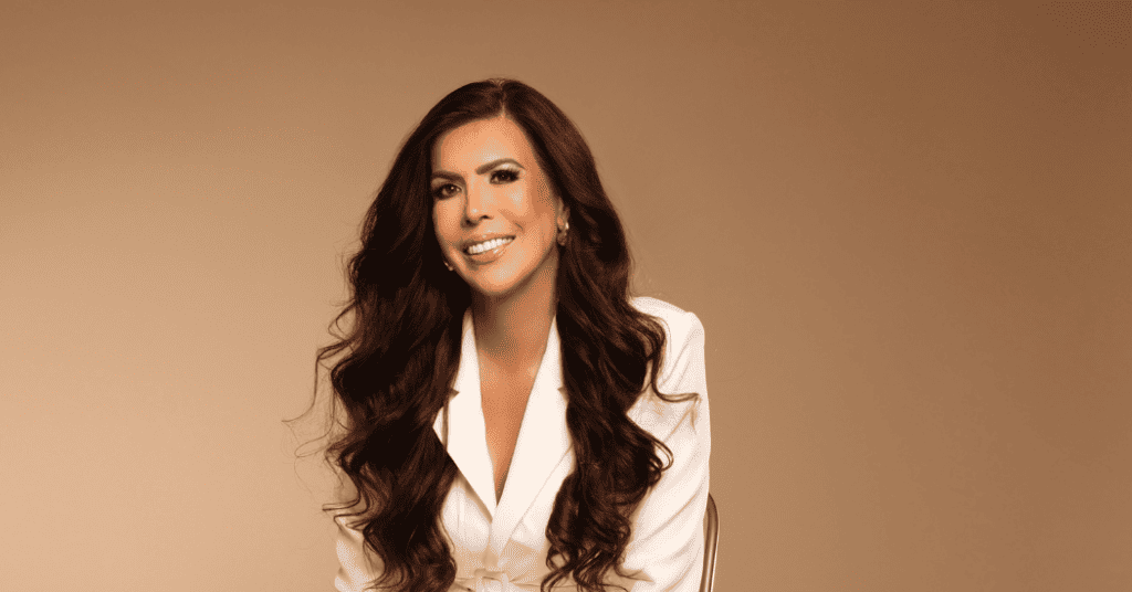 Isabel Restrepo, oradora de Miami, sonríe mientras está sentada y viste un blazer blanco sobre un fondo beige liso. Su cabello largo y rizado le da un aire seguro, lo que refleja su pasión por la mentoría en Miami.