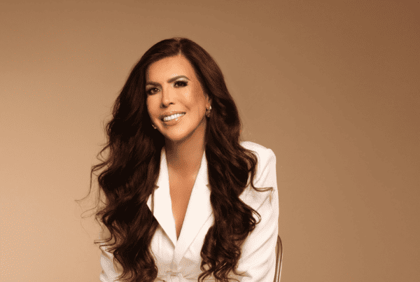 Isabel Restrepo, oradora de Miami, sonríe mientras está sentada y viste un blazer blanco sobre un fondo beige liso. Su cabello largo y rizado le da un aire seguro, lo que refleja su pasión por la mentoría en Miami.