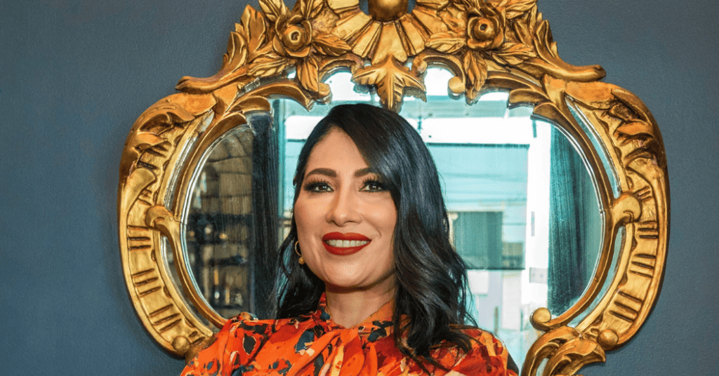 Isabel Restrepo, una destacada oradora de Miami, aparece de pie con una sonrisa frente a un espejo adornado con marco dorado; su cabello oscuro complementa su blusa con un estampado vibrante.