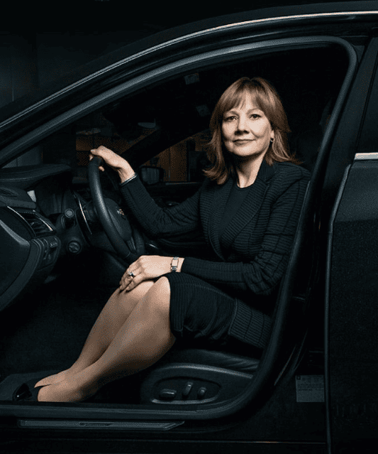 Una mujer se sienta en el asiento del conductor de un automóvil, sonriendo, con una mano en el volante y la otra apoyada en su regazo. Lleva un traje negro y tiene el pelo largo hasta los hombros. Isabel Restrepo, conocida por su mentoría en Miami, irradia confianza al volante.