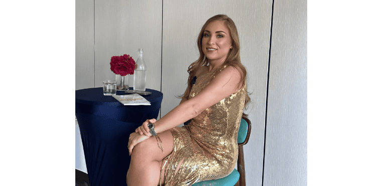 Una mujer con un vestido de lentejuelas doradas se sienta en una mesa con un mantel azul, una flor roja en un jarrón y una botella de agua etiquetada "Es Tiempo de BiYAr".