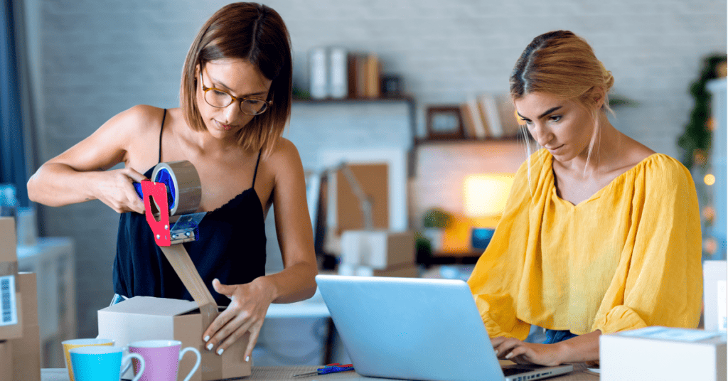 Dos mujeres trabajando; una está sellando una caja mientras la otra usa una computadora portátil. Sobre la mesa hay varias cajas, un cuaderno y tazas de colores que muestran sus habilidades de venta.