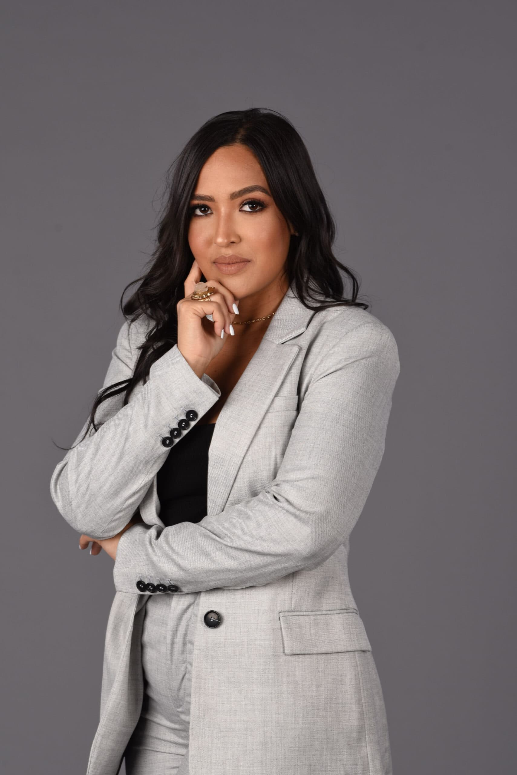 Isabel Restrepo, una persona de cabello largo y oscuro que viste un blazer y pantalones de color gris claro, está parada sobre un fondo gris y apoya la barbilla en la mano. Isabel es conocida por sus importantes contribuciones a Mentoring Miami.