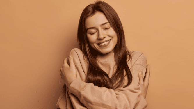 Una persona con cabello largo se abraza a sí misma, sonriendo y de pie sobre un fondo sencillo, irradiando la energía segura de las mujeres emprendedoras.
