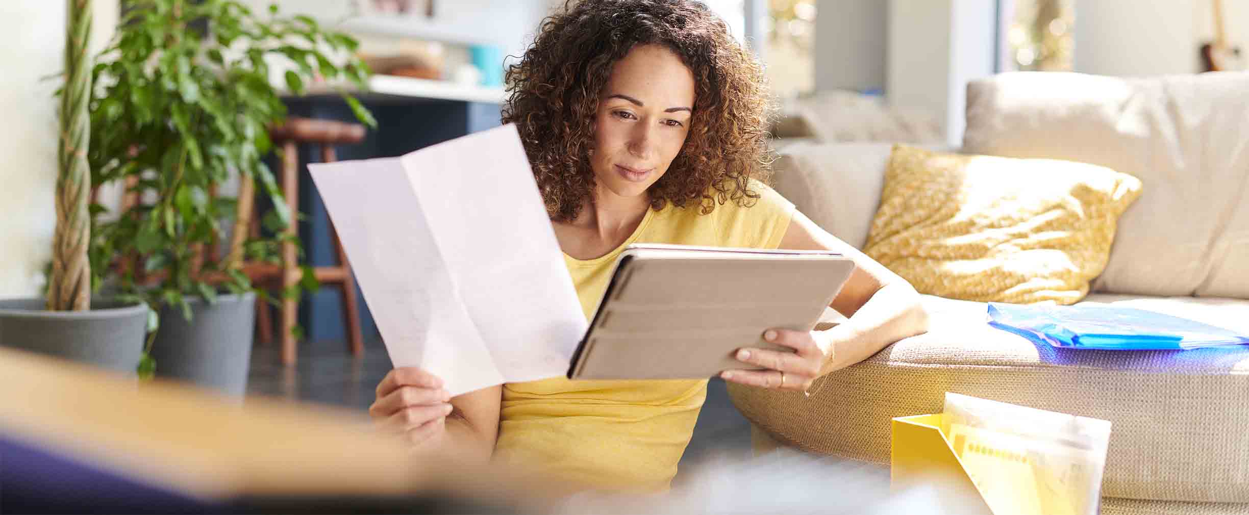 Una mujer sentada en un sofá sostiene un documento en una mano y mira una tableta en la otra. Está rodeada de artículos del hogar en una sala de estar luminosa, lo que ejemplifica la concentración y la determinación características de las mujeres exitosas.