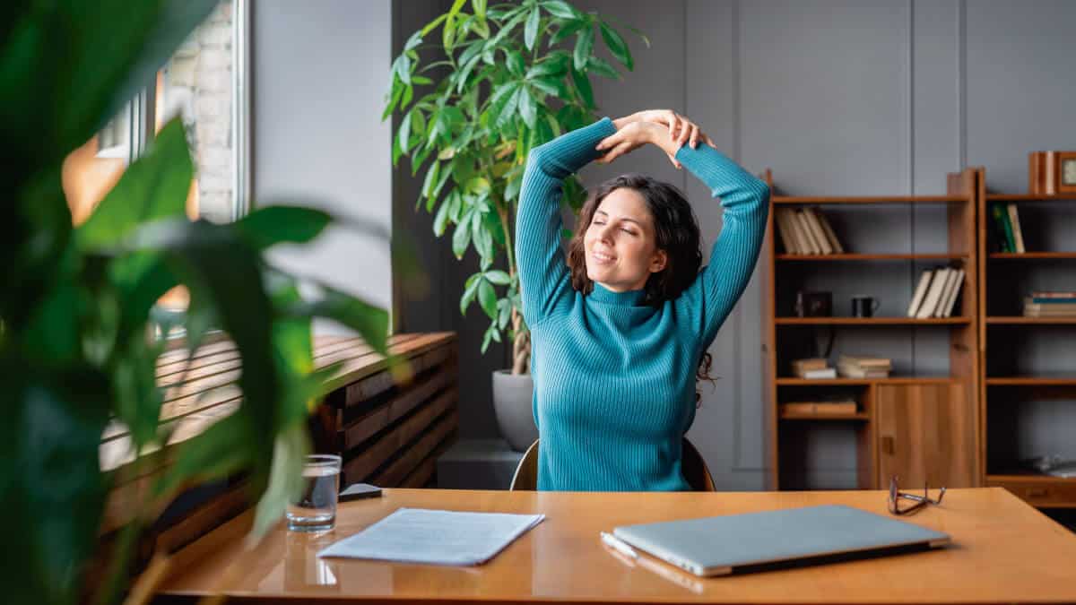 Isabel Restrepo, una reconocida oradora de Miami, se estira con los brazos sobre la cabeza en su escritorio, que tiene una computadora portátil, papeles y anteojos. Al fondo hay una gran planta y una estantería.