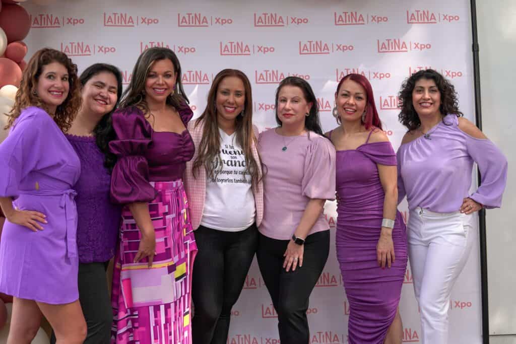 Un grupo de siete mujeres, sonrientes y posando juntas frente a un telón de fondo de la Latina Expo. Seis llevan trajes morados, mientras que una mujer en el centro, posiblemente Isabel Restrepo, lleva una camiseta blanca y pantalones negros. La escena rezuma alegría y camaradería en lo que parece un evento de Mentoring Miami.