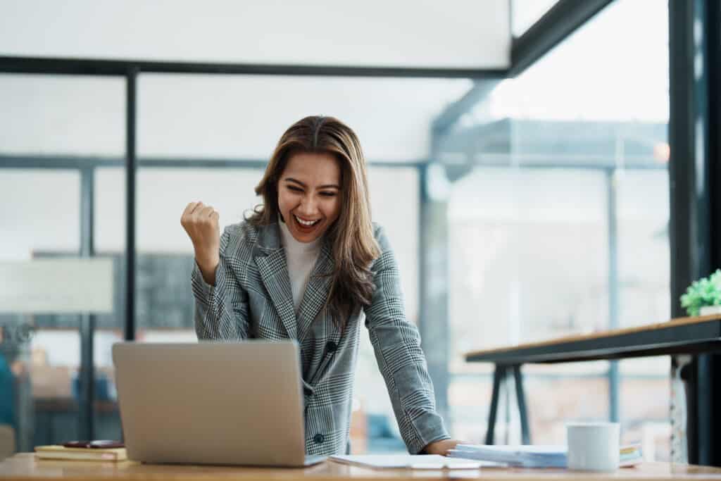 Una mujer con una chaqueta a cuadros está de pie frente a un escritorio, mirando una computadora portátil con el puño izquierdo en alto y una expresión alegre. Sobre el escritorio hay una libreta, una taza de café y una planta. Isabel Restrepo, conocida por ser mentora de los aspirantes a líderes de Miami, irradia entusiasmo mientras se prepara para su próxima sesión.