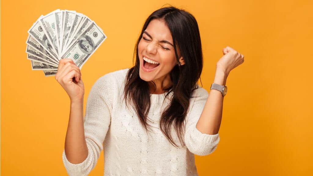 Una mujer que viste un suéter blanco celebra sosteniendo un abanico de billetes de dólares estadounidenses en una mano y levantando la otra mano en un puño contra un fondo naranja, encarnando el espíritu de las mujeres exitosas.