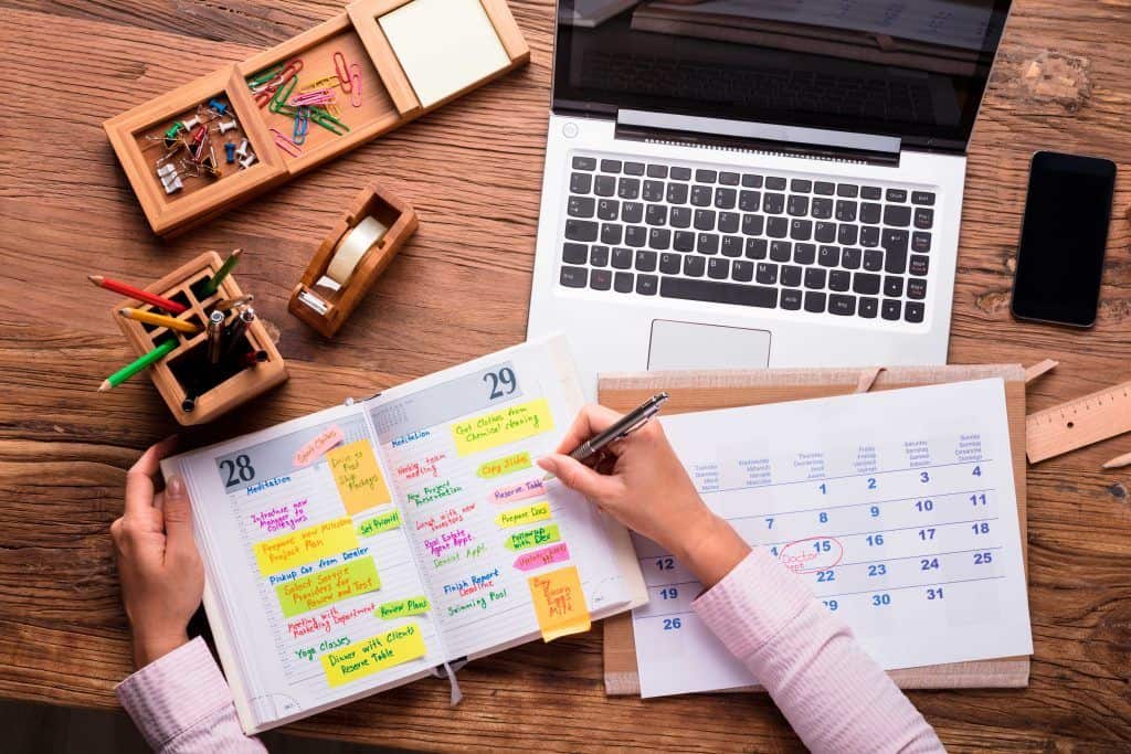 Una persona escribiendo en una agenda, con notas adhesivas de colores en el escritorio, una computadora portátil, un calendario y útiles de oficina cerca; este espacio de trabajo refleja el enfoque organizado de la oradora de Miami Isabel Restrepo mientras se prepara para su próxima sesión de tutoría en Miami.