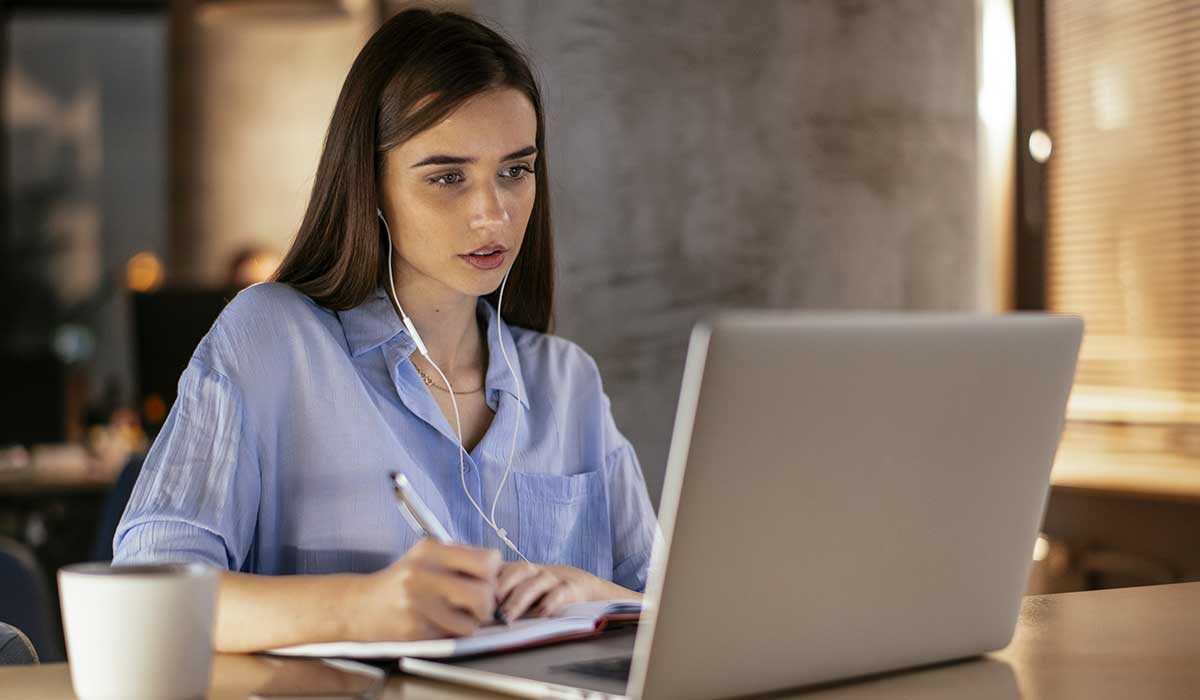 Isabel Restrepo, de pelo largo y camisa azul, está sentada en una mesa, usando una computadora portátil y tomando notas en un cuaderno. Tiene los auriculares enchufados y una taza de café con leche sobre la mesa.