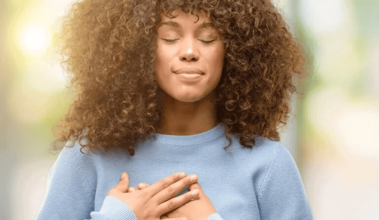 Una persona con cabello rizado y un suéter azul cierra los ojos y coloca sus manos sobre su corazón, sugiriendo un momento de reflexión o gratitud, muy parecido a Isabel Restrepo durante una de sus inspiradoras sesiones de Mentoring Miami.