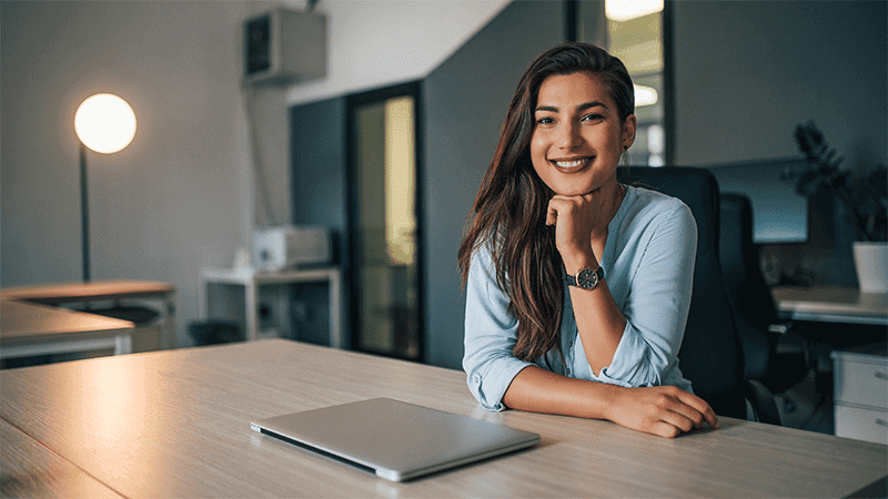Isabel Restrepo, vistiendo una camisa azul, está sentada sonriendo en un escritorio con una computadora portátil cerrada en un entorno de oficina moderno, encarnando la esencia de Mentoring Miami.