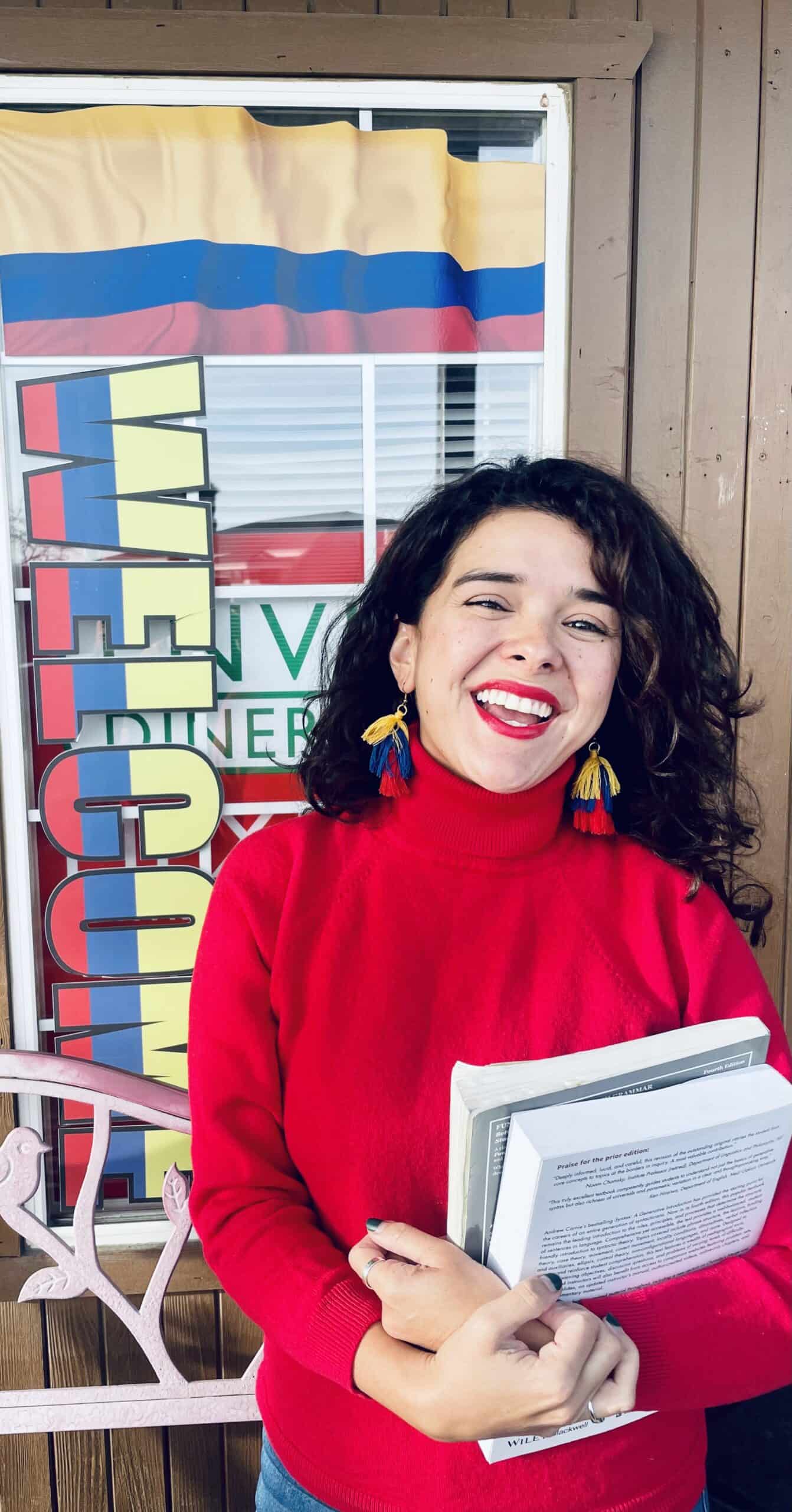 Una mujer que lleva un suéter rojo y aretes de borlas sonríe mientras sostiene unos papeles. Está parada frente a un cartel de "BIENVENIDA", que encarna la calidez y la experiencia de la reconocida oradora de Mentoring Miami, Isabel Restrepo.