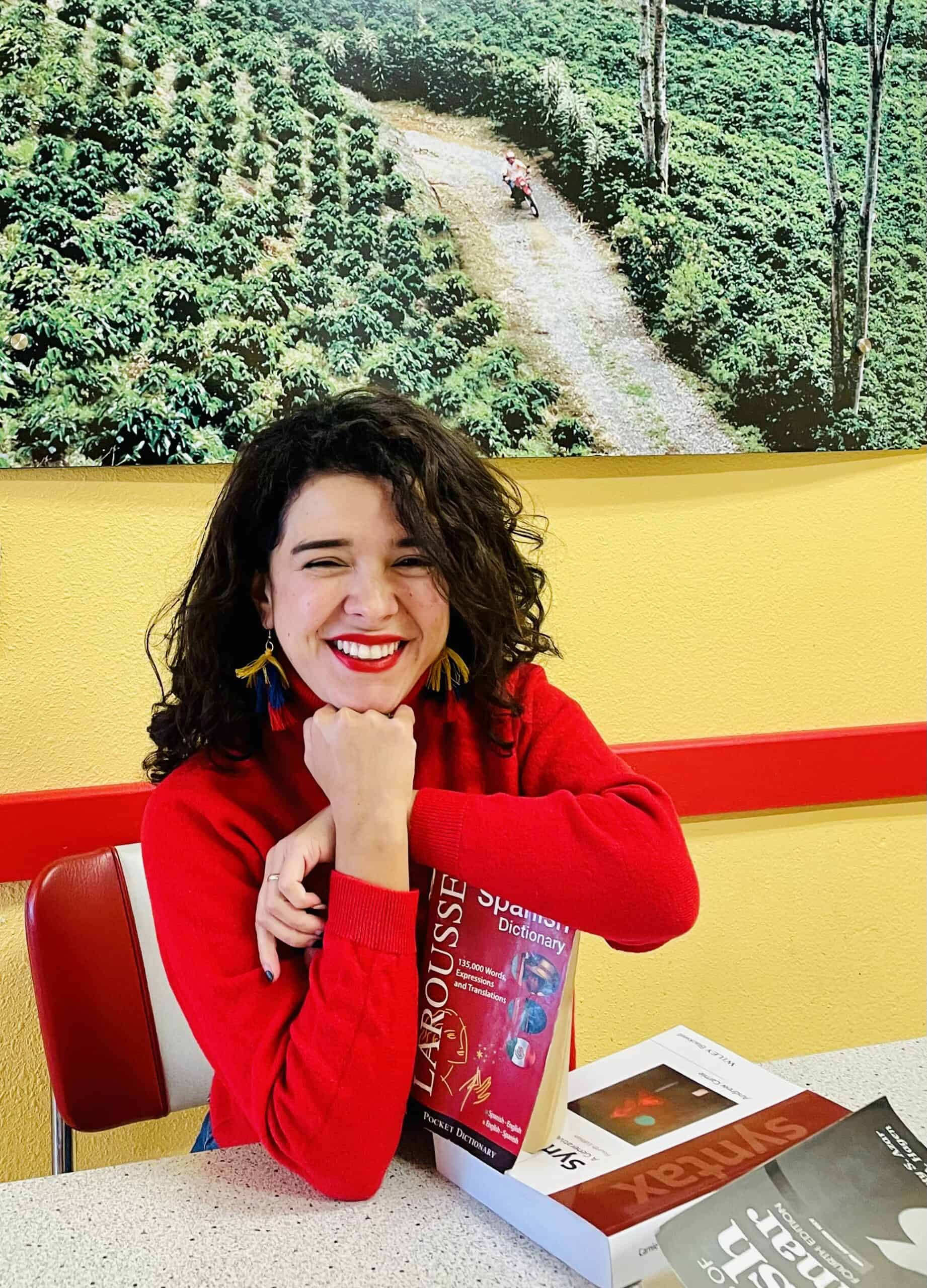 Isabel Restrepo, vestida con un suéter rojo, está sentada a una mesa, sonriendo y sosteniendo un diccionario de español. En la pared detrás de ella hay una gran fotografía de una granja. Como participante activa de Mentoring Miami, a menudo comparte su amor por el aprendizaje de idiomas con los demás.