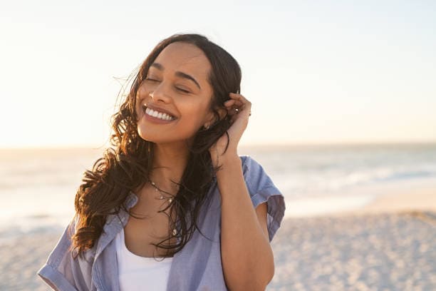 Una mujer de cabello largo y oscuro sonríe con los ojos cerrados, parada en una playa al atardecer. Se toca el cabello con una mano y viste una camisa azul claro sobre una blusa blanca, personificando la confianza serena de las mujeres emprendedoras.