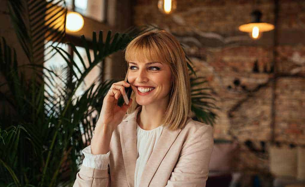 Una mujer de cabello rubio, vestida con una chaqueta clara, sonríe mientras habla por teléfono celular en un elegante ambiente interior lleno de plantas, mostrando sus habilidades de venta.