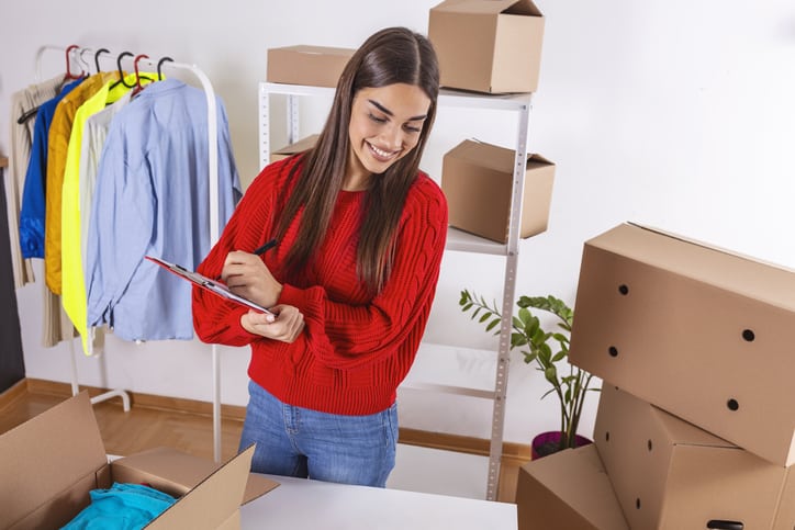 Una mujer con un suéter rojo demuestra sus habilidades de venta mientras escribe en un portapapeles mientras está de pie cerca de cajas de embalaje y ropa en un estante.