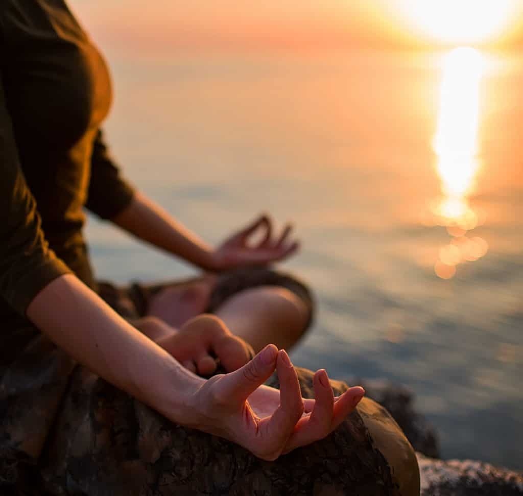 Isabel Restrepo, una reconocida oradora de Miami conocida por su trabajo como mentora, se sienta en una postura de meditación cerca de un cuerpo de agua durante el atardecer. La serena luz del atardecer baña la escena, reflejando su actitud tranquila y su dedicación a la atención plena.