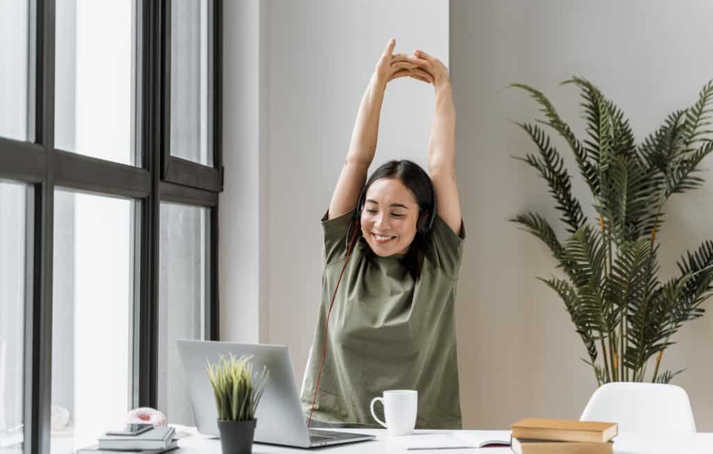 Una mujer con auriculares estira los brazos por encima de la cabeza mientras está sentada en un escritorio con una computadora portátil, una taza de café y algunos libros. Se ve una planta al fondo, junto a una gran ventana. Las sesiones de mentoría en Miami suelen comenzar de esta manera para Isabel Restrepo.