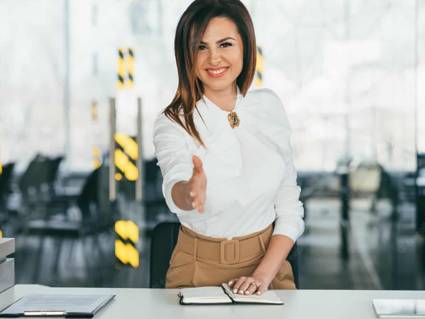 Una mujer sonriente con blusa blanca y pantalón beige, posiblemente Isabel Restrepo, extiende su mano para un apretón de manos en un entorno de oficina, ejemplificando el espíritu acogedor de Mentoring Miami.
