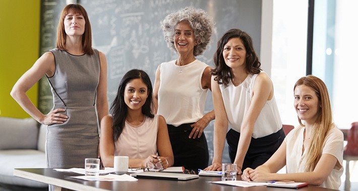 Un grupo de cinco mujeres vestidas con ropa profesional se reúnen alrededor de una mesa de conferencias, sonriendo a la cámara. Entre ellas se encuentra Isabel Restrepo, una reconocida oradora de Miami, conocida por sus inspiradoras sesiones de mentoría en Miami.