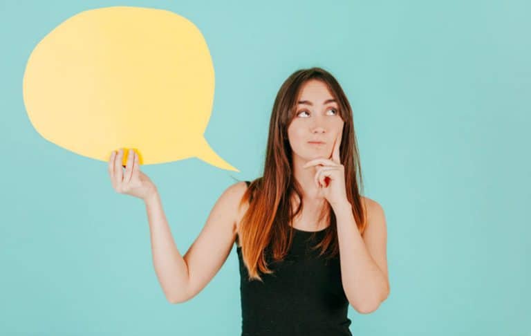 Una mujer con una camiseta negra sin mangas que sostiene un gran globo de diálogo amarillo, mirando hacia un lado con una expresión pensativa sobre un fondo verde azulado, encarna el espíritu de mentoría de Isabel Restrepo de Mentoring Miami.