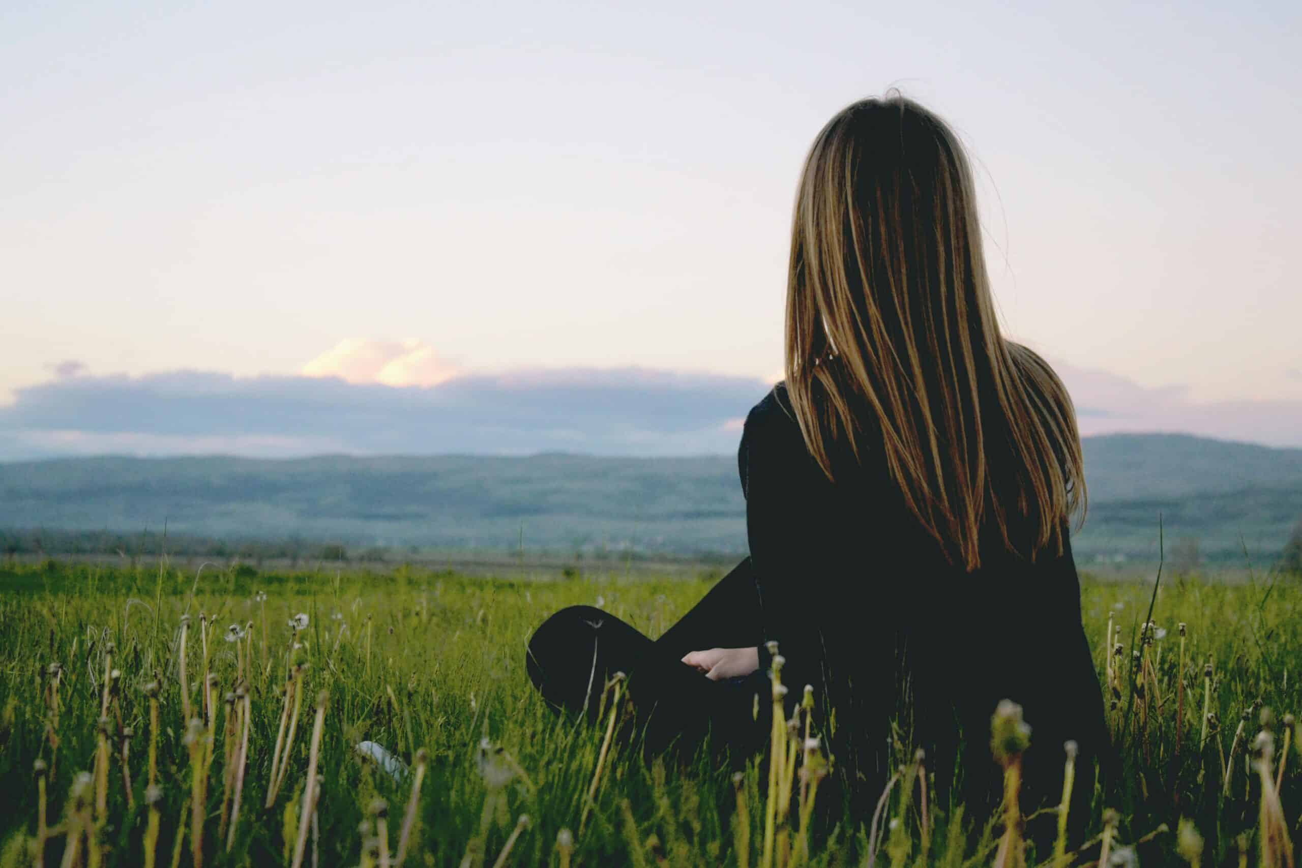 Una persona con cabello largo se sienta en un campo de hierba, mirando hacia un paisaje distante bajo un cielo crepuscular, que recuerda los momentos serenos de Isabel Restrepo cuando no está dando clases en Miami.