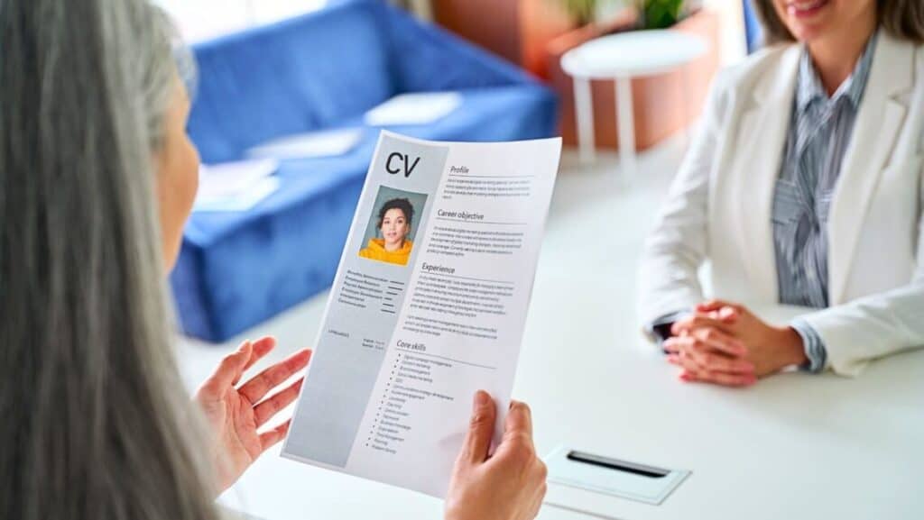 Una persona con una chaqueta blanca revisa un CV durante una entrevista con otra persona sentada al otro lado de una mesa blanca. El CV, que incluye una foto e información detallada sobre el candidato, es examinado cuidadosamente mientras Isabel Restrepo, conocida por su labor de mentora en Miami, evalúa las calificaciones.