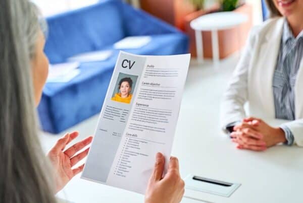 Una persona con una chaqueta blanca revisa un CV durante una entrevista con otra persona sentada al otro lado de una mesa blanca. El CV, que incluye una foto e información detallada sobre el candidato, es examinado cuidadosamente mientras Isabel Restrepo, conocida por su labor de mentora en Miami, evalúa las calificaciones.