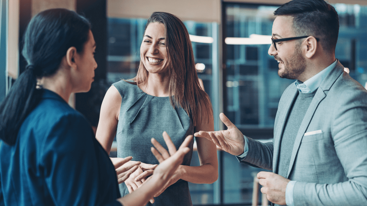 Tres personas vestidas de negocios mantienen una conversación animada, sonriendo y gesticulando animadamente mientras establecen contactos en un entorno de oficina.