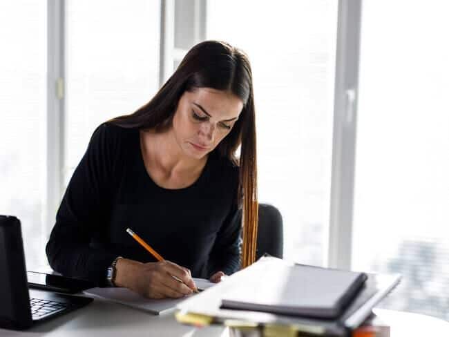 Una mujer está sentada en un escritorio, concentrada en escribir en un cuaderno con un lápiz. Junto a ella hay una computadora portátil y una pila de papeles, posiblemente relacionados con sus esfuerzos de networking.