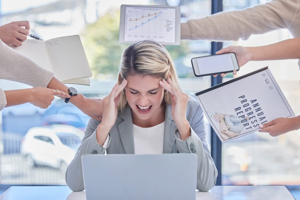 Una mujer se sienta en un escritorio con una computadora portátil, sosteniendo su cabeza en señal de frustración mientras múltiples manos a su alrededor presentan varios dispositivos y documentos.