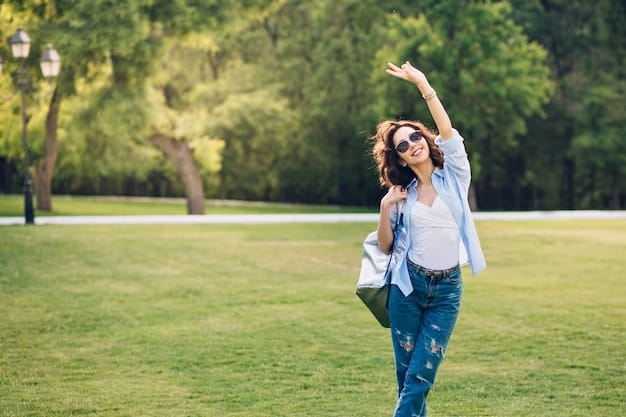 Una mujer con gafas de sol saluda mientras camina por un parque. Lleva una camiseta azul claro sobre una camiseta blanca, unos vaqueros azules y lleva un bolso grande. Al fondo se ven árboles y una farola que capturan la esencia de disfrutar de la vida al aire libre.