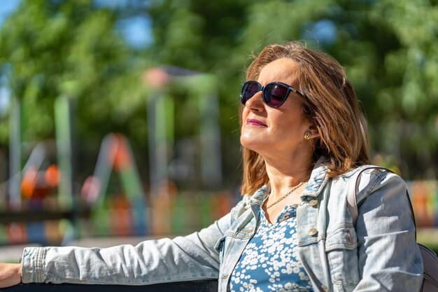 Una mujer con gafas de sol y una chaqueta vaquera está sentada al aire libre, disfrutando de la luz del sol con los ojos cerrados. En el fondo borroso, un parque infantil contribuye a la atmósfera serena de este momento de aire libre.